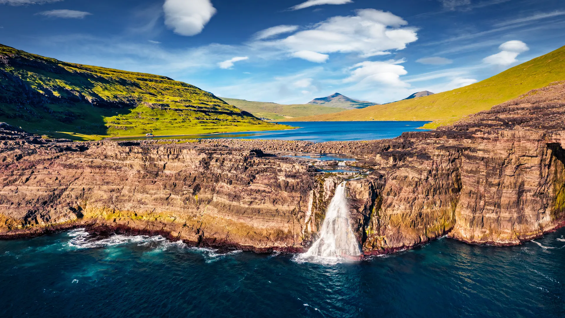 Bøsdalafossur waterfall flowing into the ocean in the Faroe Islands, with steep cliff faces surrounding the waterfall.