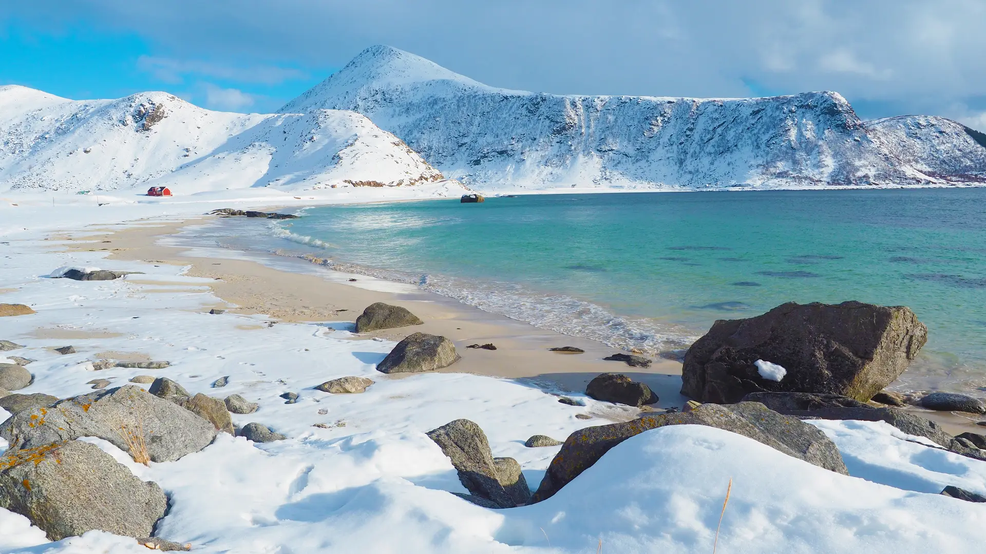 Winter shoreline at Haukland Beach in the Lofoten Islands, Norway, showing flat coastal terrain beneath snow-covered mountains.